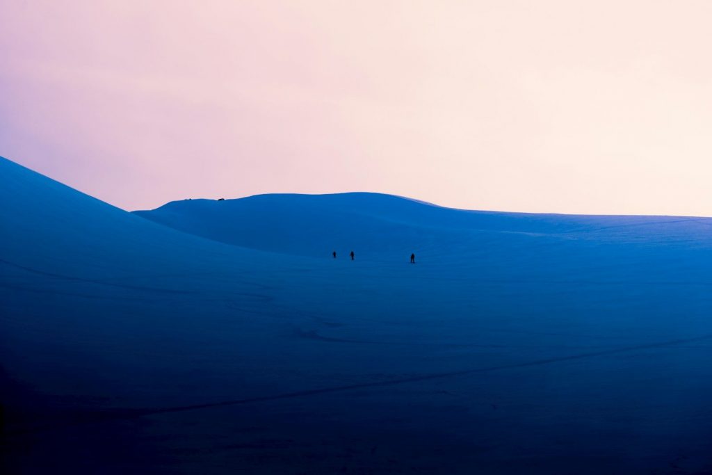 People walking across vast blue dunes under a pink sky.
